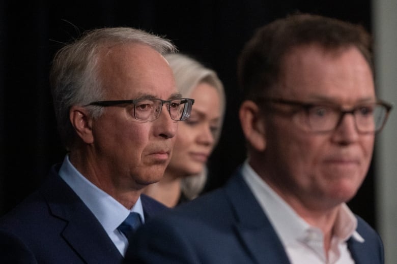 A head and shoulders shot of Falcon and Rustad. Falcon is out of focus in the front, with the sharp Focus on Rustad just behind him. Falcon wears a suit over a white open-collared shirt, while Rustad wears a dark blue suit over a pale blue shirt and a dark blue tie.