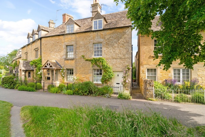 A two-storey stone cottage, part of a curving terrace, on a country road. It has a wooden porch and two dormer windows in the roof