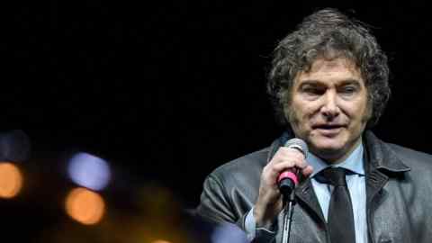 A man with curly hair and sideburns, wearing a black leather jacket and tie, speaks into a microphone against a dark background