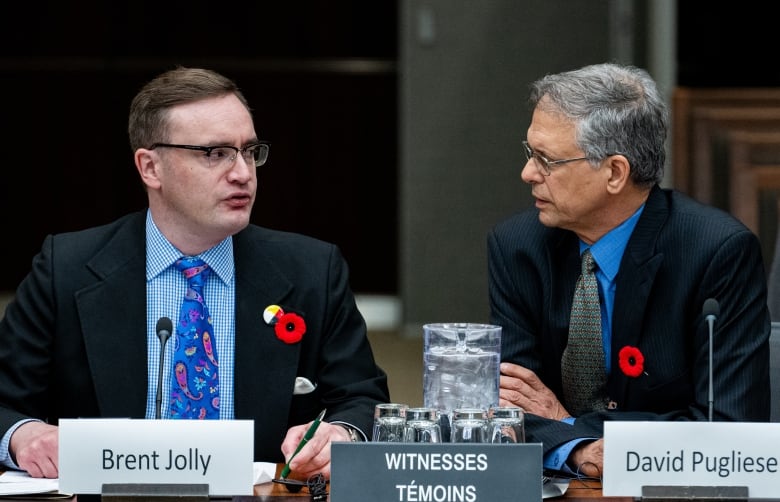 Two men in suits sit at a desk and speak to each other.