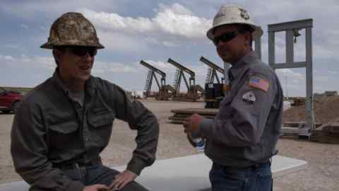 Two oil workers wearing hard hats and protective clothing take a break near oil pump jacks