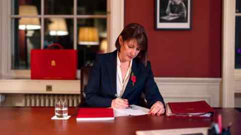 Rachel Reeves is seated at her desk in No 11 Downing Street, preparing for the Autumn Budget 2024. She wears a dark suit with a poppy pin and is writing on documents. A red briefcase is visible on the windowsill behind her. A portrait of Ellen Wilkinson hangs on the wall.