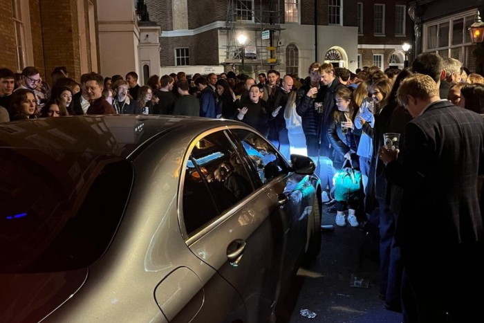 A large crowd of people stands tightly packed outside, holding drinks and socializing on a dimly lit street. A car is stopped with its headlights illuminating the crowd, attempting to move through but blocked by the gathering. 