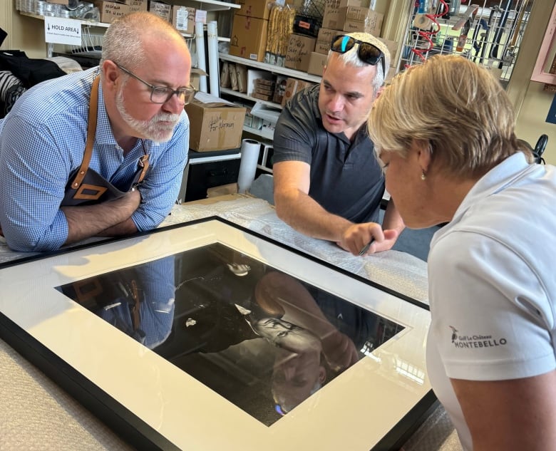 Three people lean over to look at a framed portrait under glass.