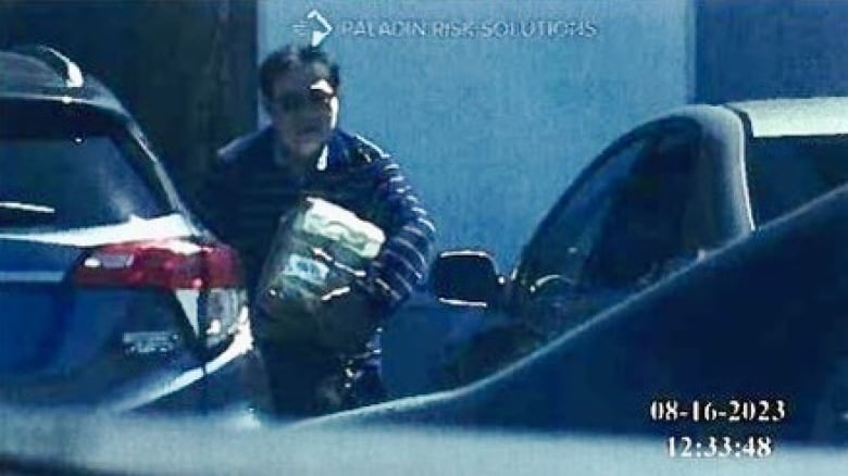 A man in a grainy video walks between two cars holding a number of small packages wrapped in clear plastic.