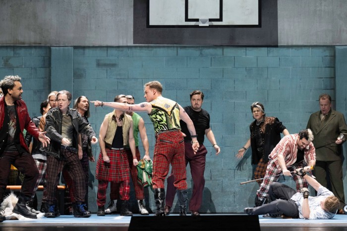 In a fight scene from an opera, in a functional space, a group of people wearing punk-style tartan gather aggressively; a man gestures with his arm, while to one side a man wielding a golf club is wrestled to the ground