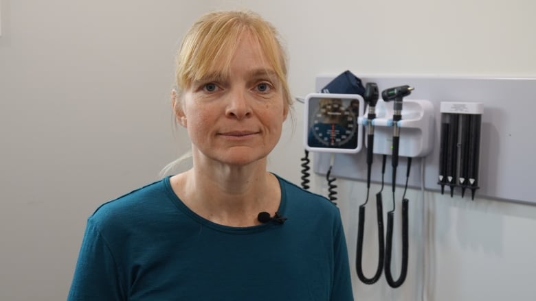 A woman with blonde hair and a blue shirt stands in a white room with medical equipment, including a blood pressure gauge and otoscopes from examining a patient's ears, hanging on the wall behind her.