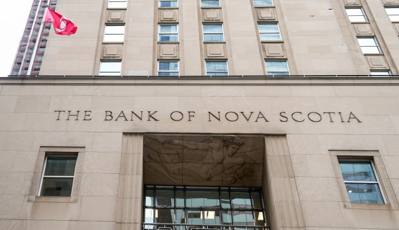 An image of a building taken from the perspective of someone standing on the street and looking up at it. The building is beige and says THE BANK OF NOVA SCOTIA on it, with a Canadian flag on the top. 