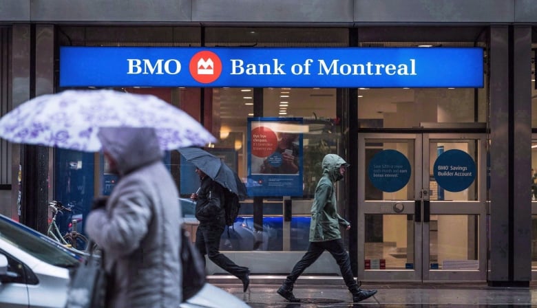 People walk past a building that says BMO BANK OF MONTREAL. Is it raining and most are wearing raincoats or holding umbrellas. 