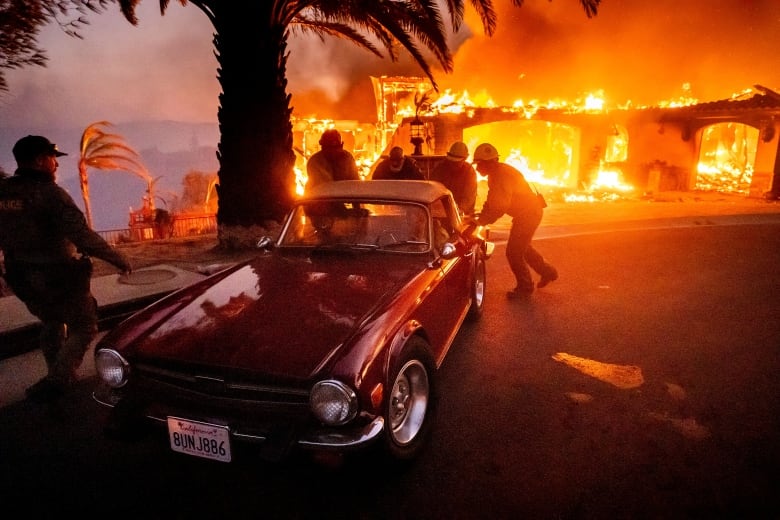 A vintage sports car is pushed by firefighters in the foreground as a burning structure and palm trees are shown in the background.