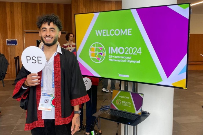 Samed AlHajajla stands smiling in front of a screen displaying &quot;Welcome IMO 2024, 65th International Mathematical Olympiad.&quot; He holds a sign labeled &quot;PSE&quot;  and is wearing a traditional embroidered garment.