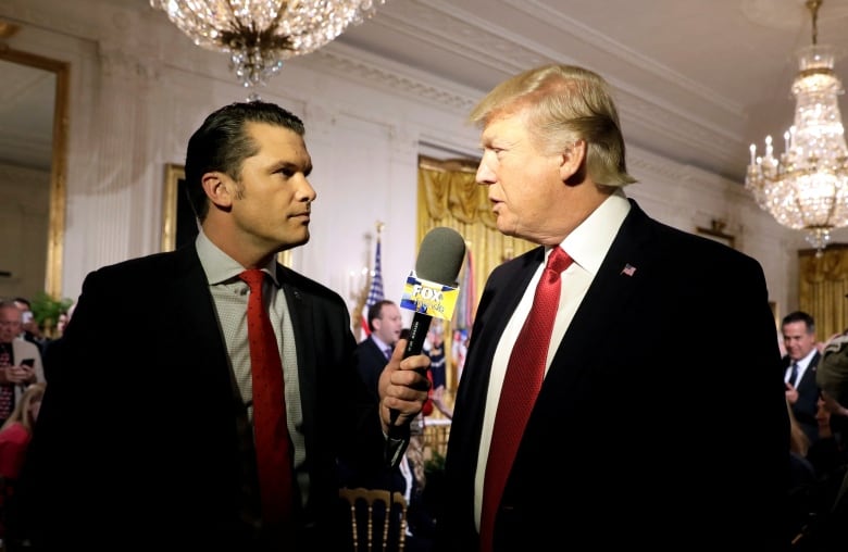 A dark haired, clean shaven man in a suit and tie holds a microphone near the face of an older, clean shaven light-haired man in a suit and tie who is speaking.