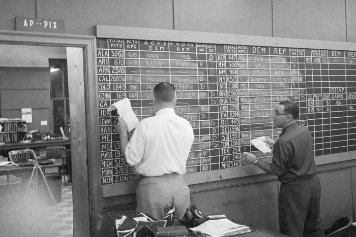 Associated Press staffers are seen updating a large chalkboard with election results. The board displays totals for various states, categorising votes for different parties. 