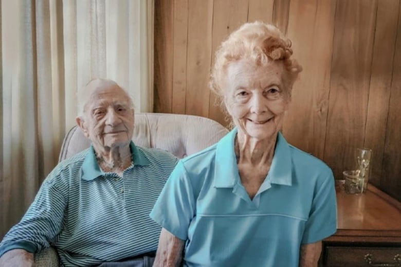 A elderly couple, both dressed in light blue, pose for a photo side by side in front of a wood-paneled wall. 