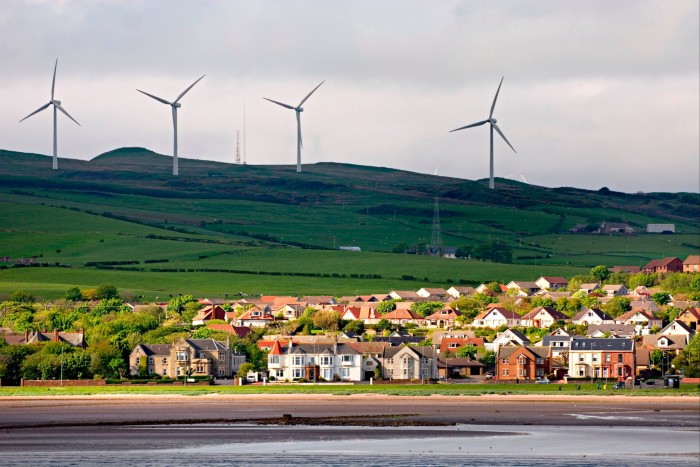 A small coastal town, photographed from the sea, with a view of wind turbines on the green hills beyond 