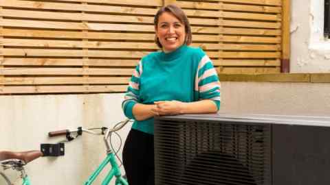 A woman in a blue jumper with striped sleeves leans on a heat pump, which is a large black rectangular object with what looks like a fan at the front