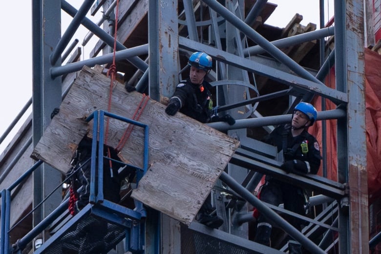 Two men wearing blue helmets are dwarfed by the scaffolding they're on. One man grips a large piece of wood with one hand.