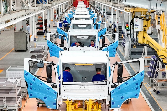 An assembly line in a factory with multiple white vehicle cabs lined up, each with doors open and technicians in blue uniforms working inside